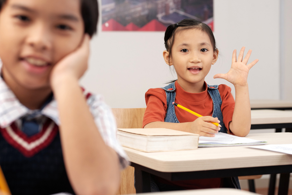Asian pupils sitting in classroom with worksheets and girl putting up hand to answer