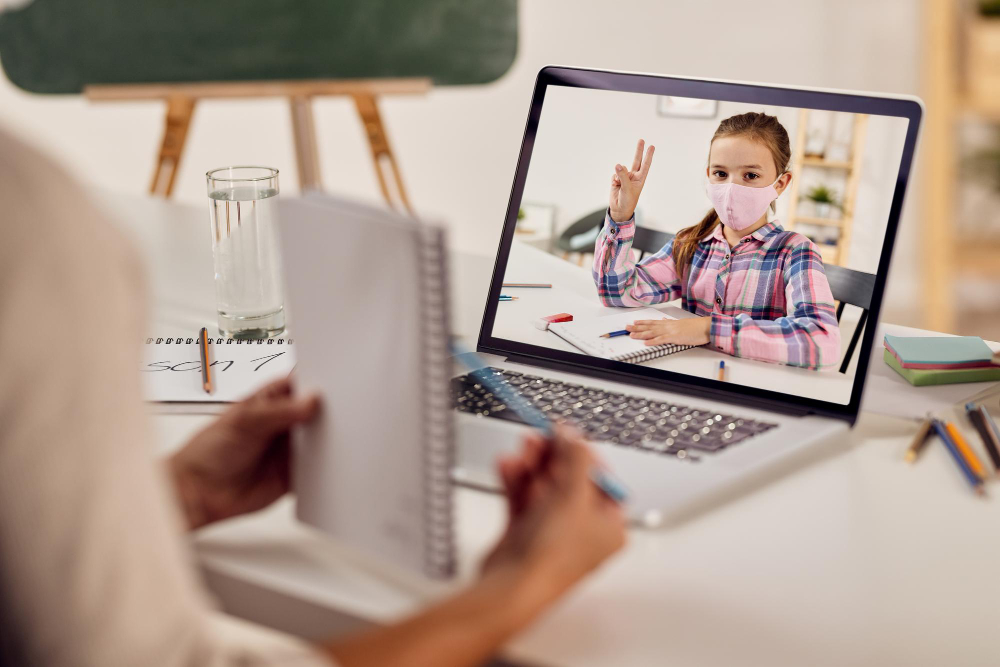 A child wearing a pink mask is smiling and giving a peace sign during a virtual learning session on a laptop, while an adult holds a notebook and pen.